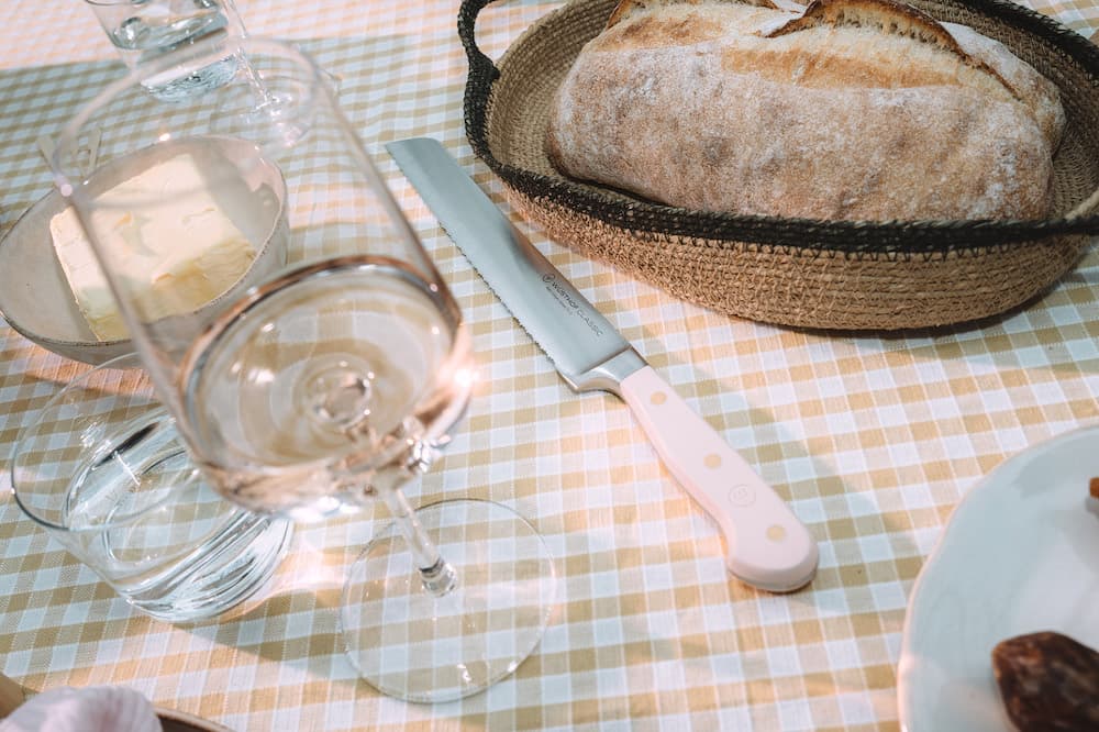 A table setting with a loaf of bread in a basket, a knife, a glass of water, and a glass of wine on a checkered tablecloth.