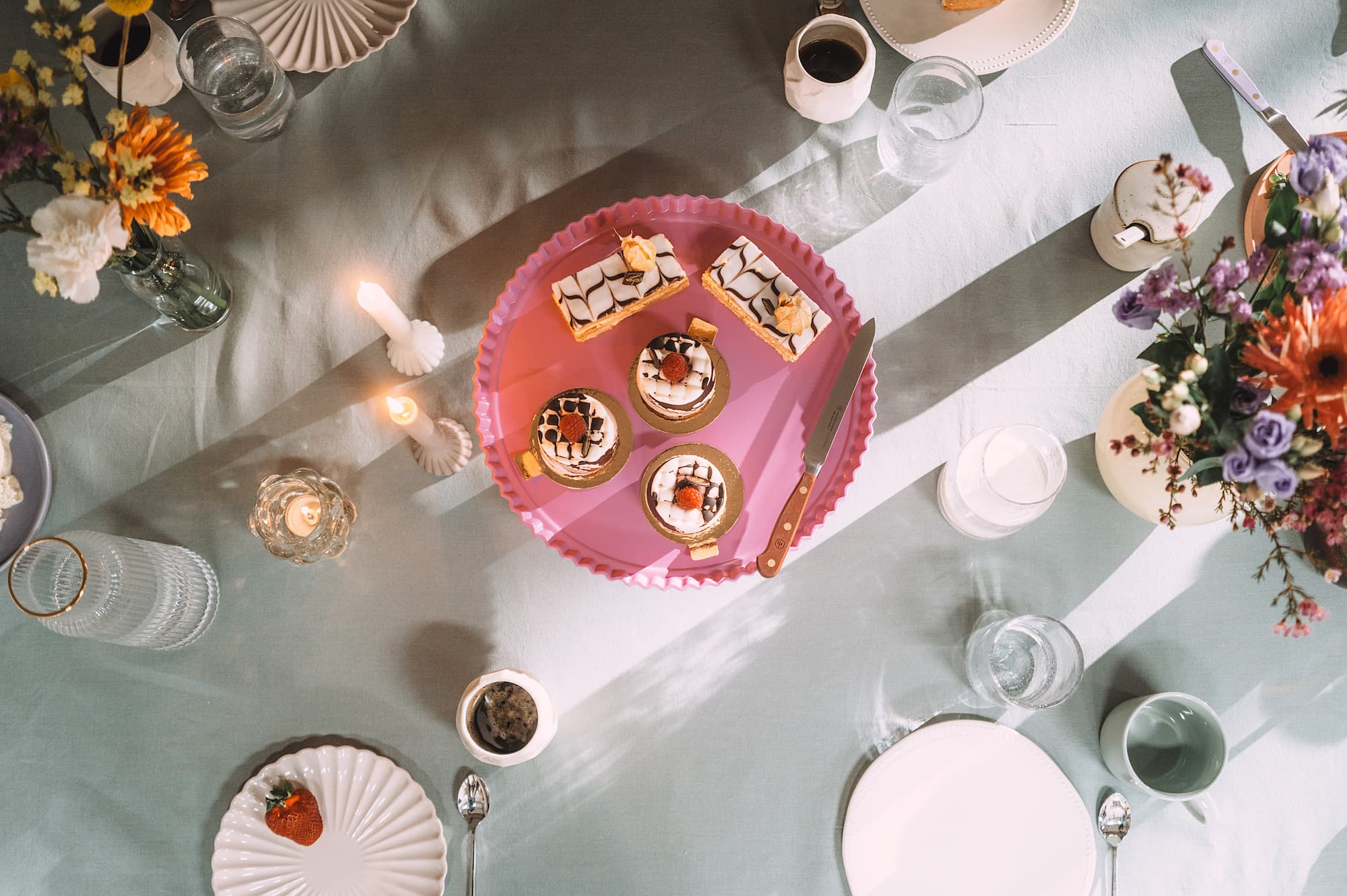 A table set with desserts on a pink tray, surrounded by flowers, candles, plates, and glasses on a light tablecloth.