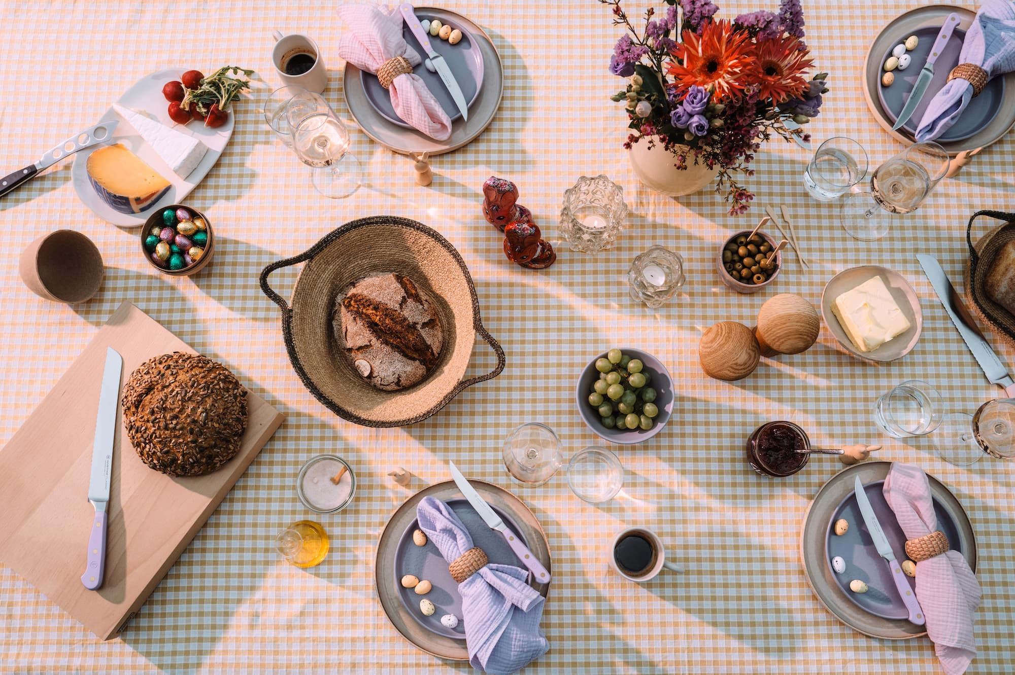 Aerial view of a picnic table with bread, cheese, olives, flowers, and neatly arranged plates with napkins on a checkered tablecloth.