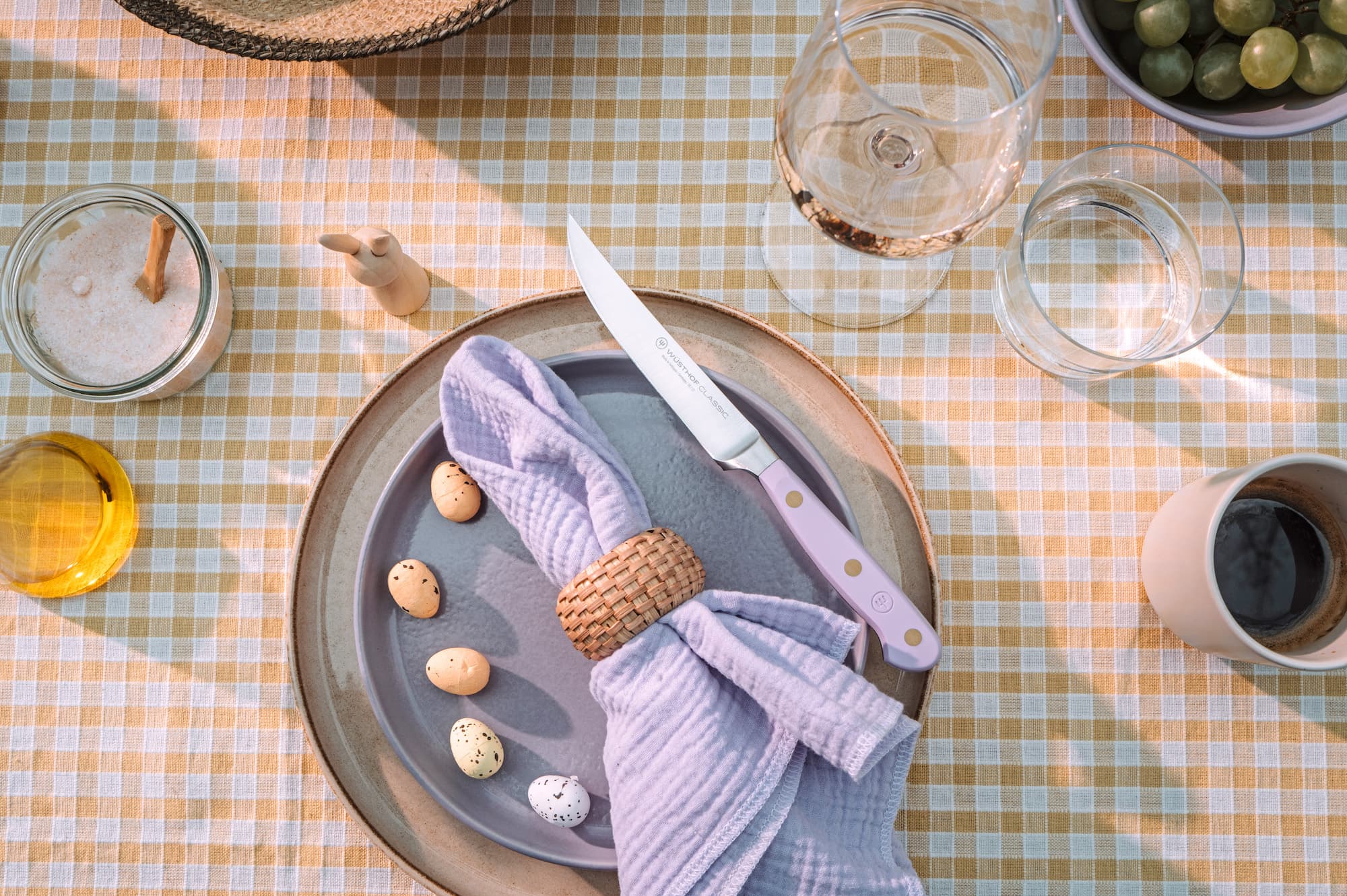 A table setting with a gray plate, speckled eggs, knife, purple napkin, glasses, and a cup on a checkered tablecloth.
