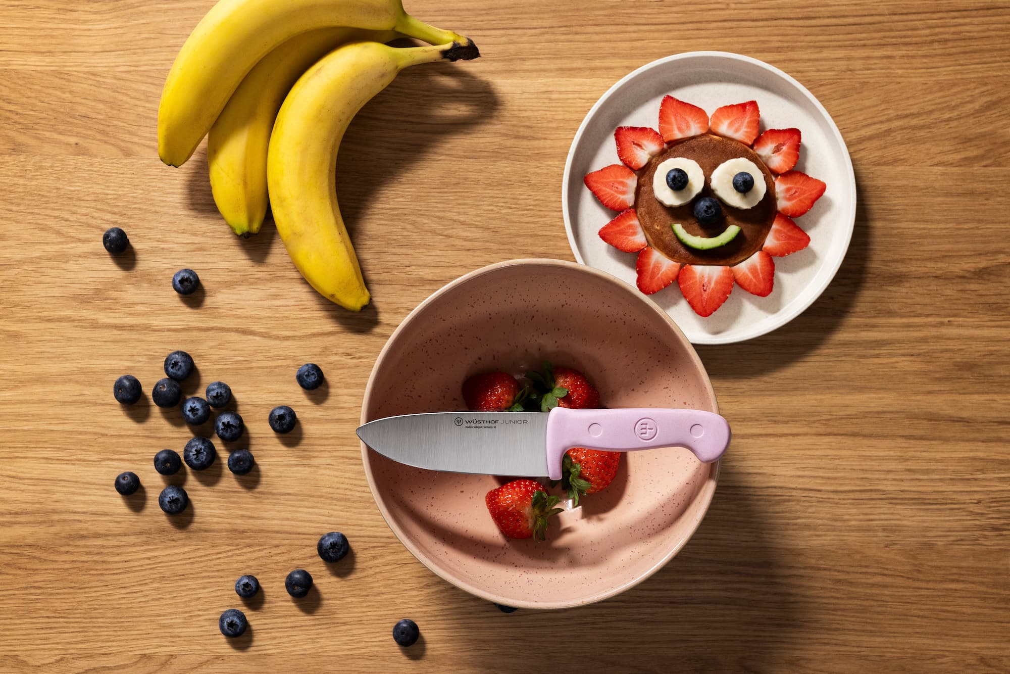 pink young kids knife on top of a fruit bowl next to bananas, blueberries and a happy face made of fruit