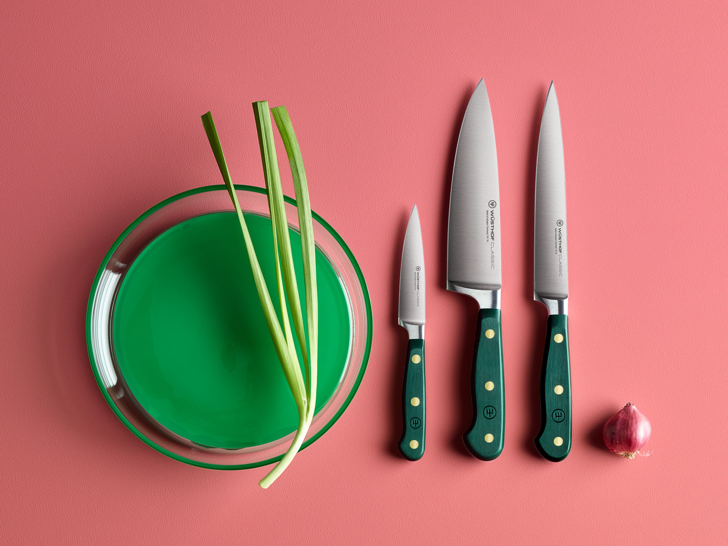 A glass bowl with green liquid and green onions, three knives with green handles, and a small red onion on a pink surface.