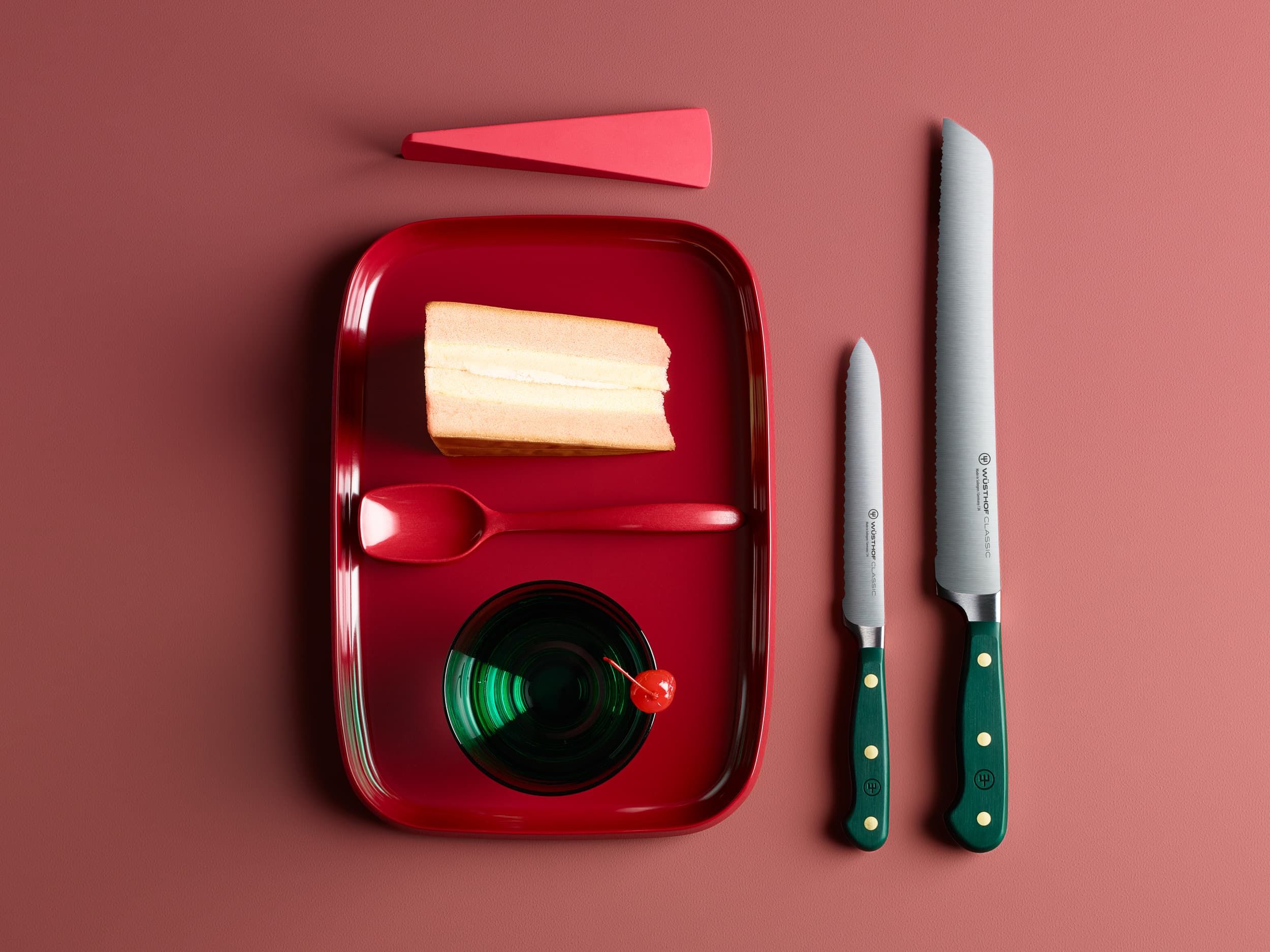 A slice of cake on a red tray with a spoon, a green glass with a cherry, a red triangle, and two knives on a red background.
