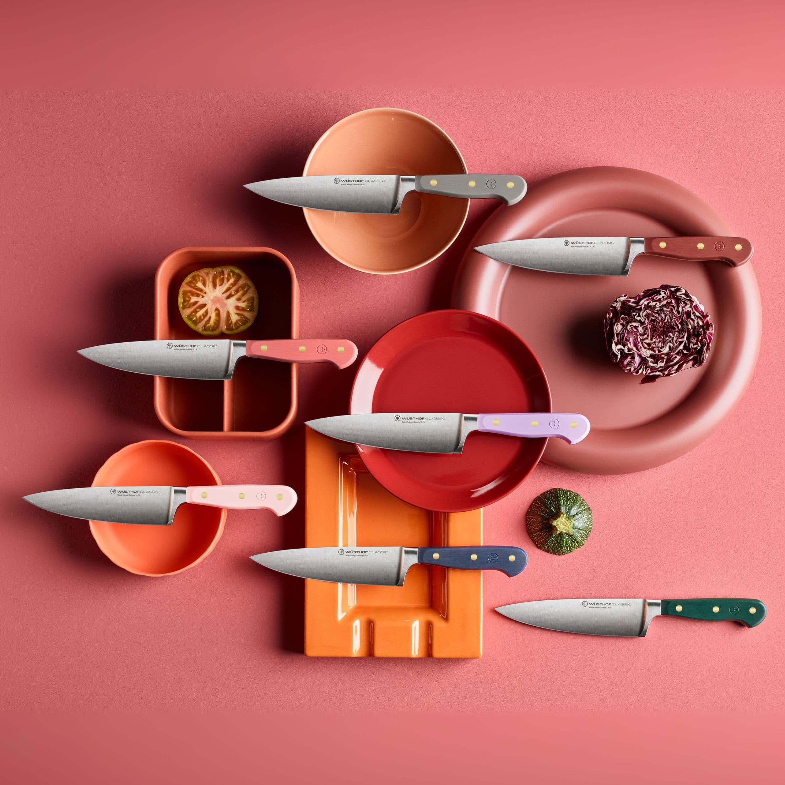A colorful arrangement of kitchen knives with various handles, placed on vibrant plates and cutting boards against a pink background.
