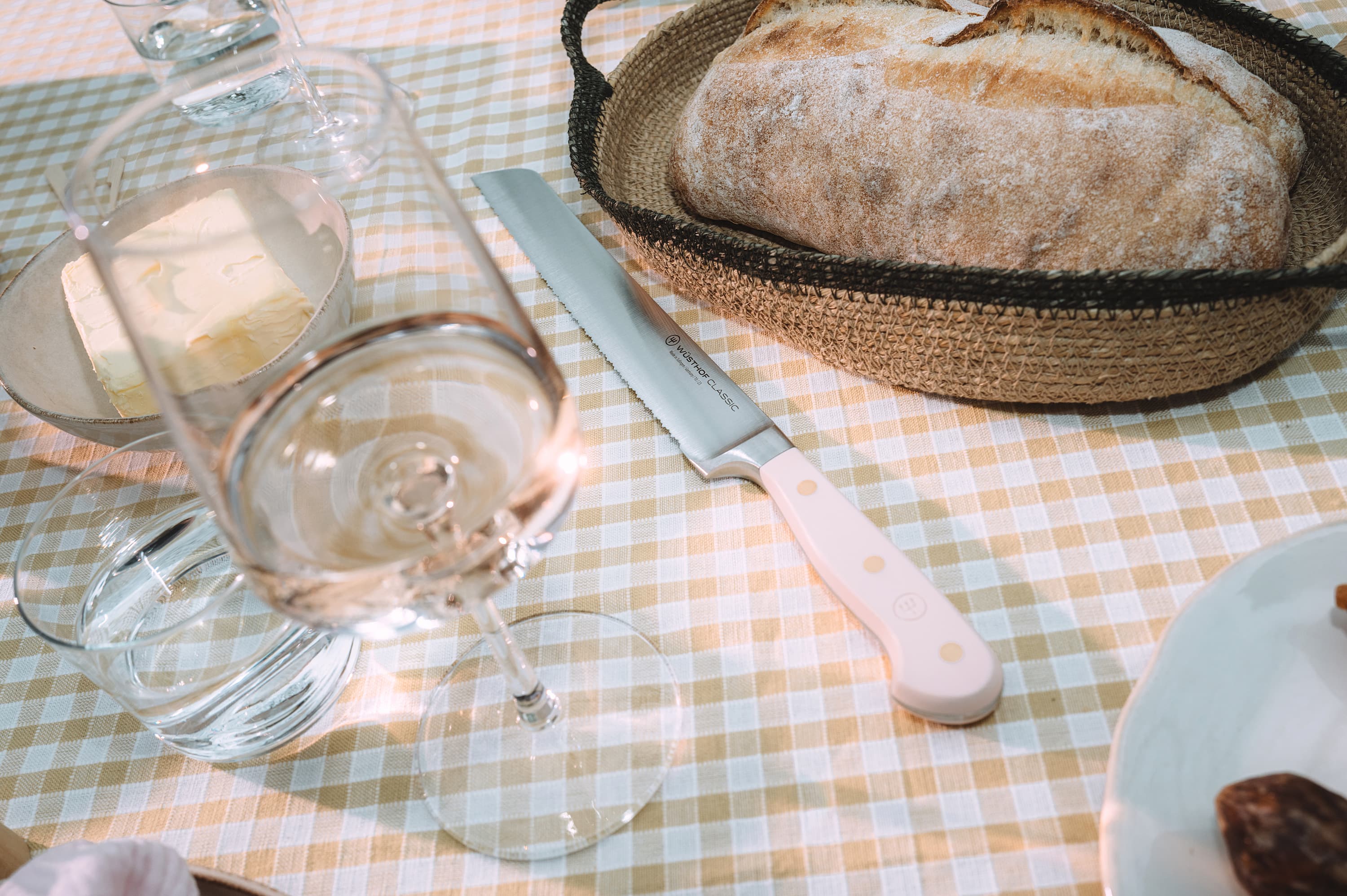 A table setting with a loaf of bread in a basket, a bread knife, a wine glass, and a dish of butter on a checkered tablecloth.