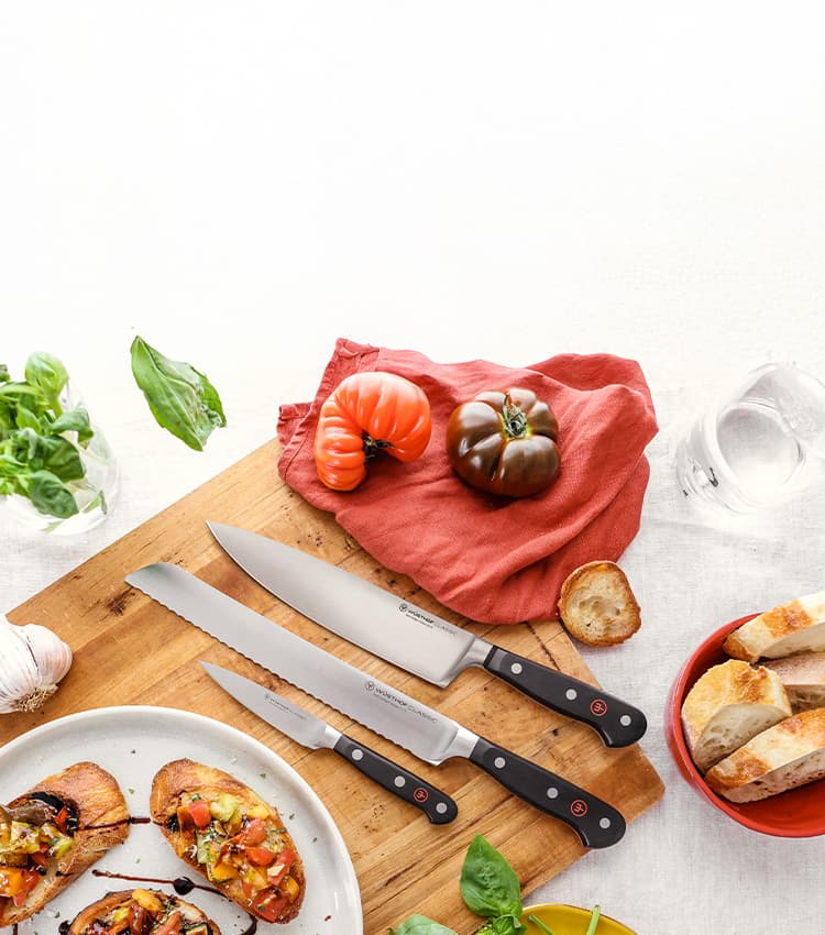 Wooden cutting board with three knives, heirloom tomatoes, basil, and bread. Bruschetta on a plate nearby.